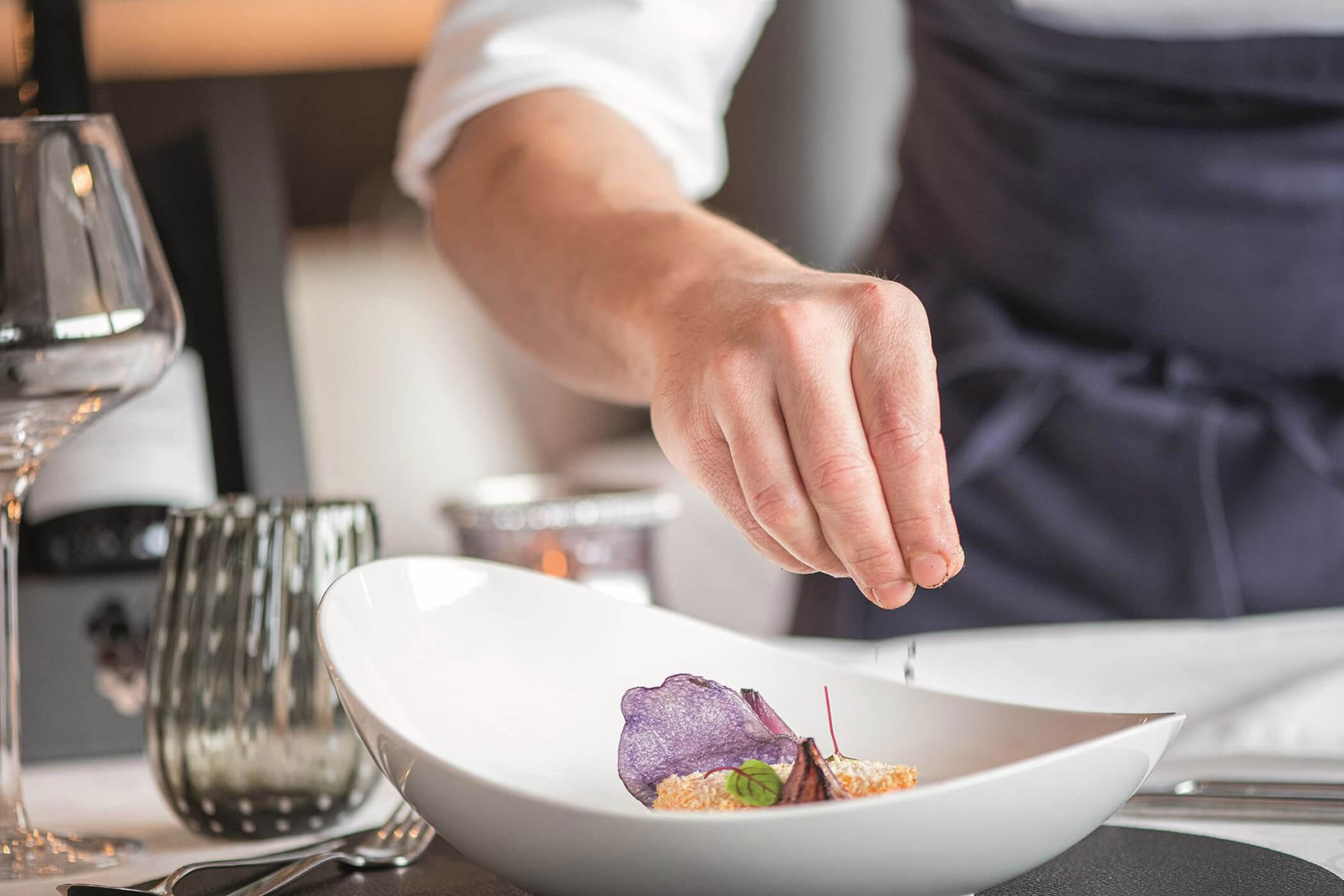 Ein Koch garniert ein angerichtetes Gericht mit einem violetten Gemüsechip. - Hotel DAS ROYAL