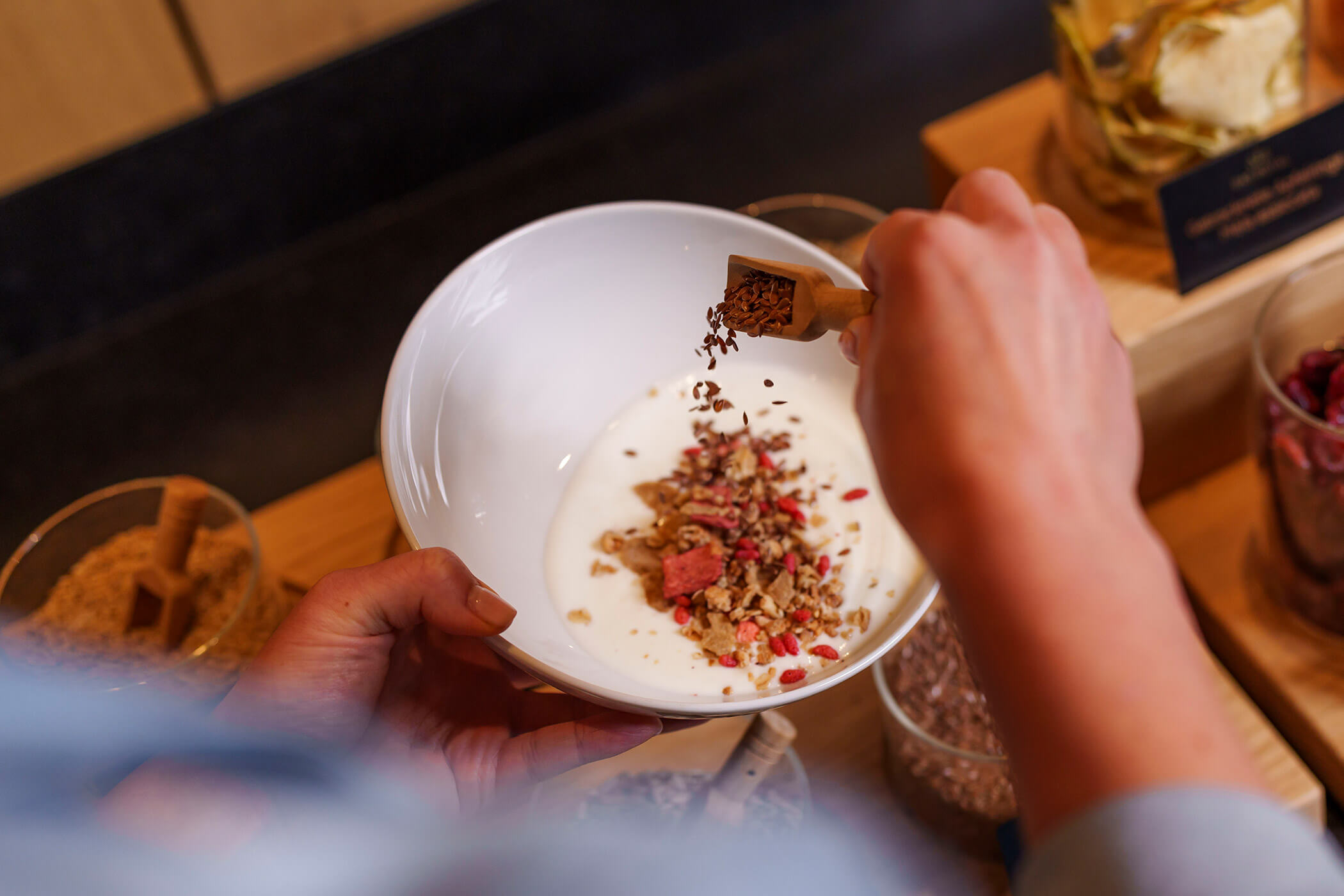 Jemand streut mit einer Holzschaufel Samen in eine Schüssel mit Joghurt und Müsli. - Hotel DAS ROYAL