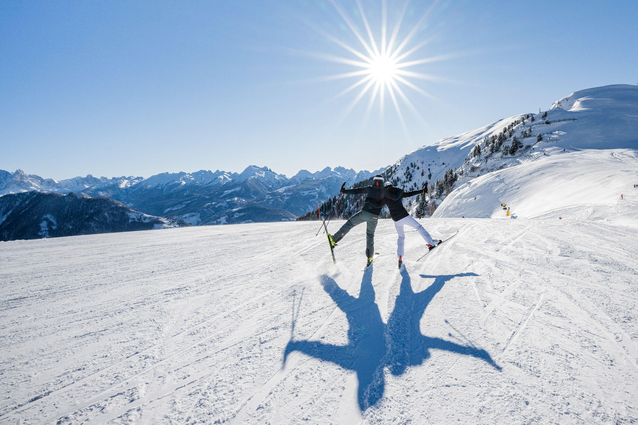 Zwei Skifahrer springen unter strahlendem Sonnenschein auf einem verschneiten Berghang. - Hotel DAS ROYAL