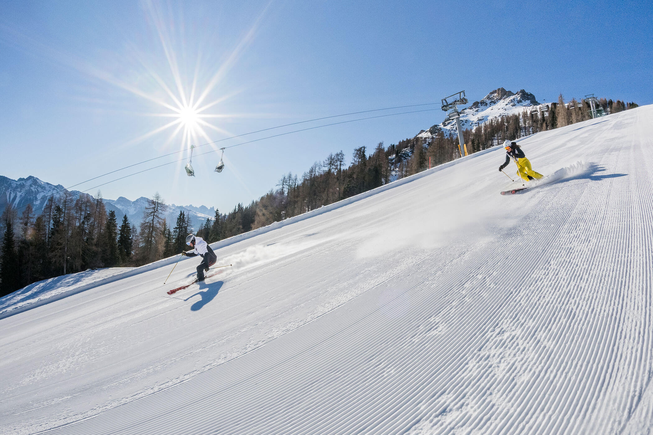 Zwei Skifahrer, die unter strahlendem Sonnenschein einen präparierten Berghang hinunterfahren. - Hotel DAS ROYAL