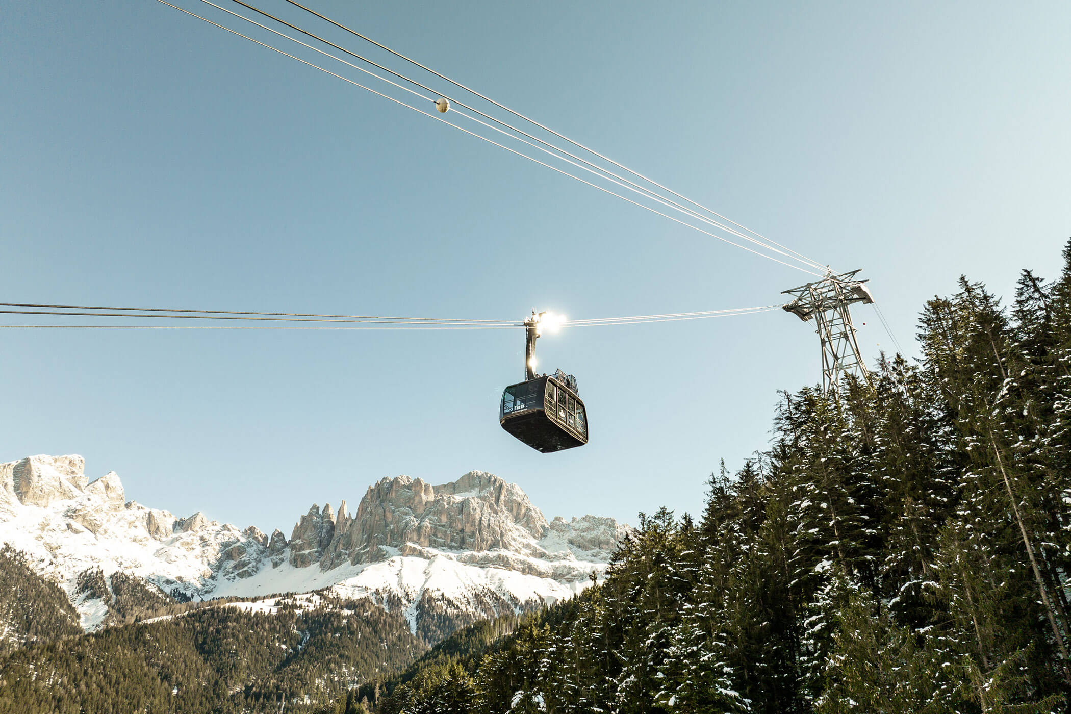 Eine Seilbahn fährt über schneebedeckte Gipfel und Kiefern hinweg auf einen Bergturm zu. - Hotel DAS ROYAL